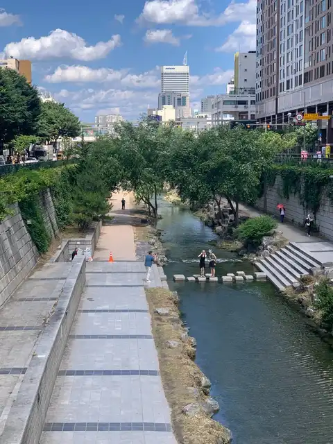 Cheonggyecheon Stream Weitblick – erhöhter Weg und Trittsteine mit den Wolkenkratzern des Zentrums von Seoul hinter der Baumreihe