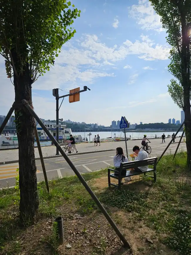Rad- und Laufweg im Han-River-Park – Pärchen auf einer Bank im Vordergrund, Radfahrer und Fußgänger am Ufer, Seoul-Skyline auf der anderen Flussseite