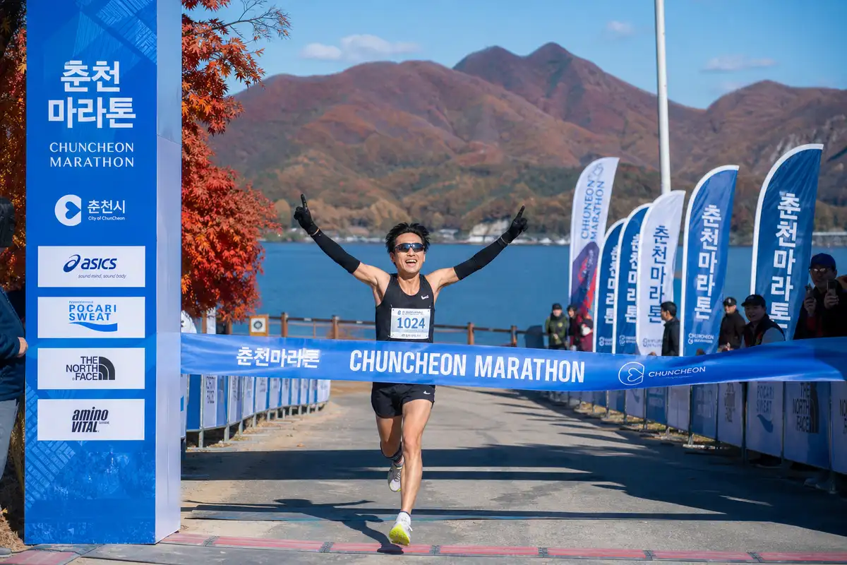 Runner crossing finish line in Chuncheon with autumn foliage and Uiam Lake visible — Korea marathon 2026
