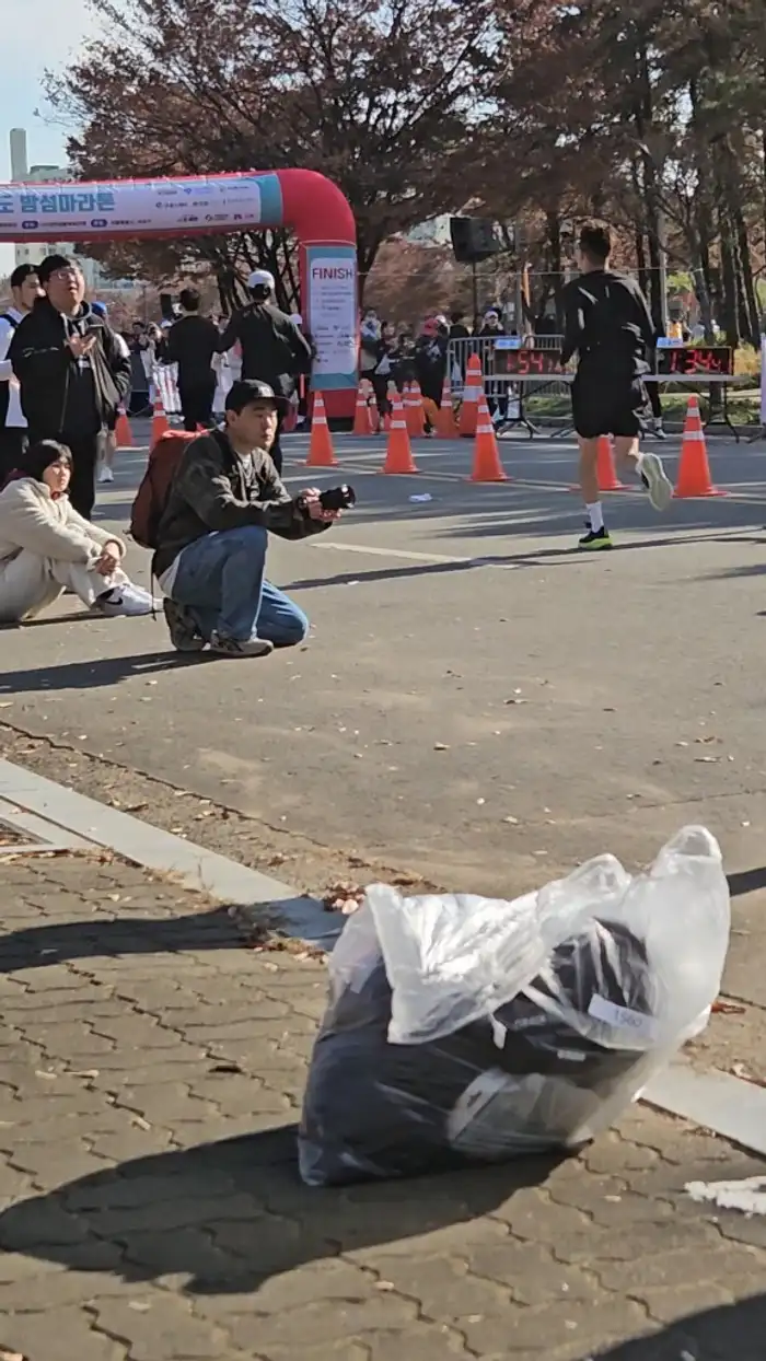 Korean autumn marathon finish line — runners crossing the finish with timer board visible, spectators and photographers at the line