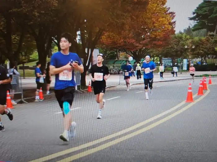 Runners with race bibs in an autumn Korean park — orange and red foliage lining the race course, morning light