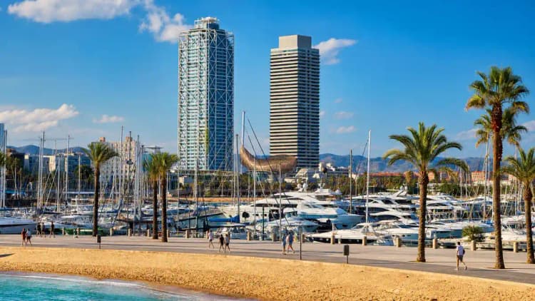 Port Olímpic marina in Barcelona by day — yachts moored along a palm-lined promenade with the beach and waterfront hotels near Casino Barcelona