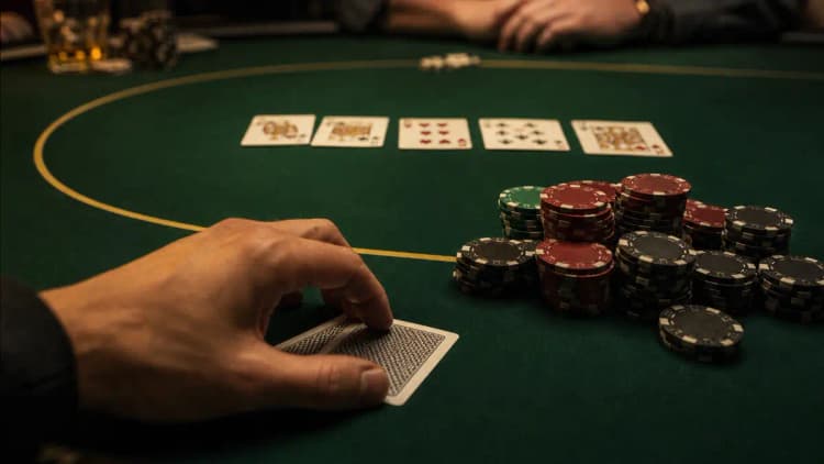 Ein volles Fünf-Karten-Board auf dem grünen Filz neben einem großen Chipstapel, während ein Spieler zwei verdeckte Karten hält und abwägt, ob er auf einer späteren Street folden soll