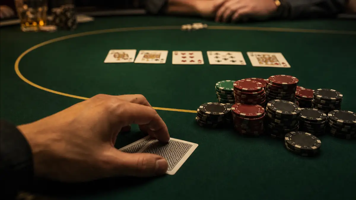 A full five-card board on the green felt beside a large pile of chips as a player holds two face-down cards, weighing whether to fold on a later street